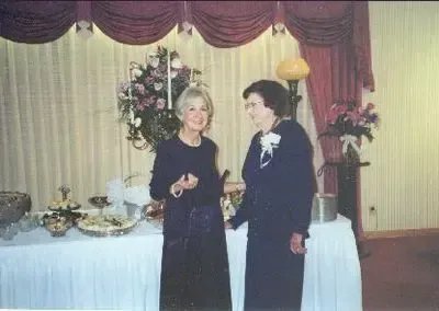 Two women standing by a buffet table, possibly at an event. One holds an item, smiles; the other wears a corsage.