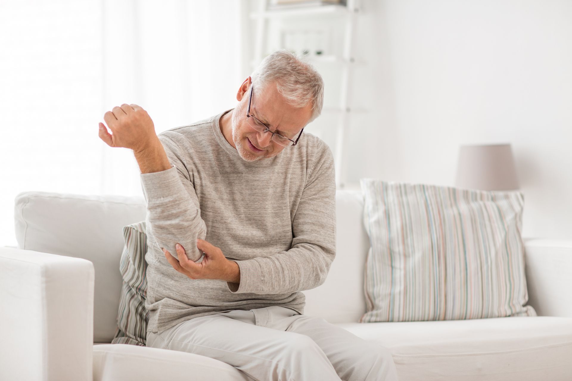 An elderly man is sitting on a couch holding his elbow in pain.