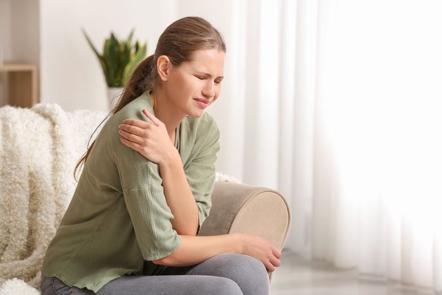 A woman is sitting on a couch holding her shoulder in pain.