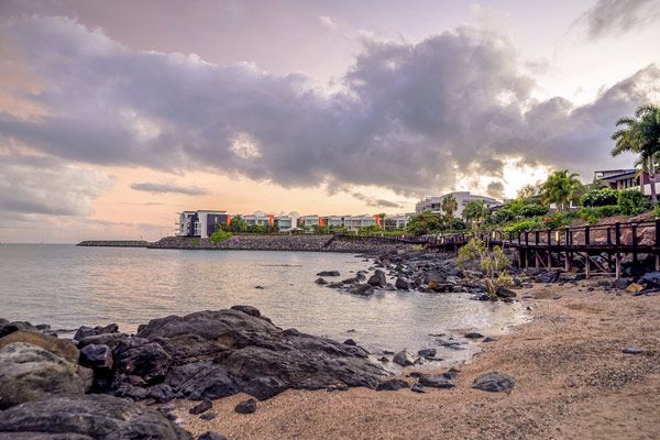 A Beach With Rocks and Buildings on the Shore at Sunset — Coastal Arborist Services in Whitsundays, QLD