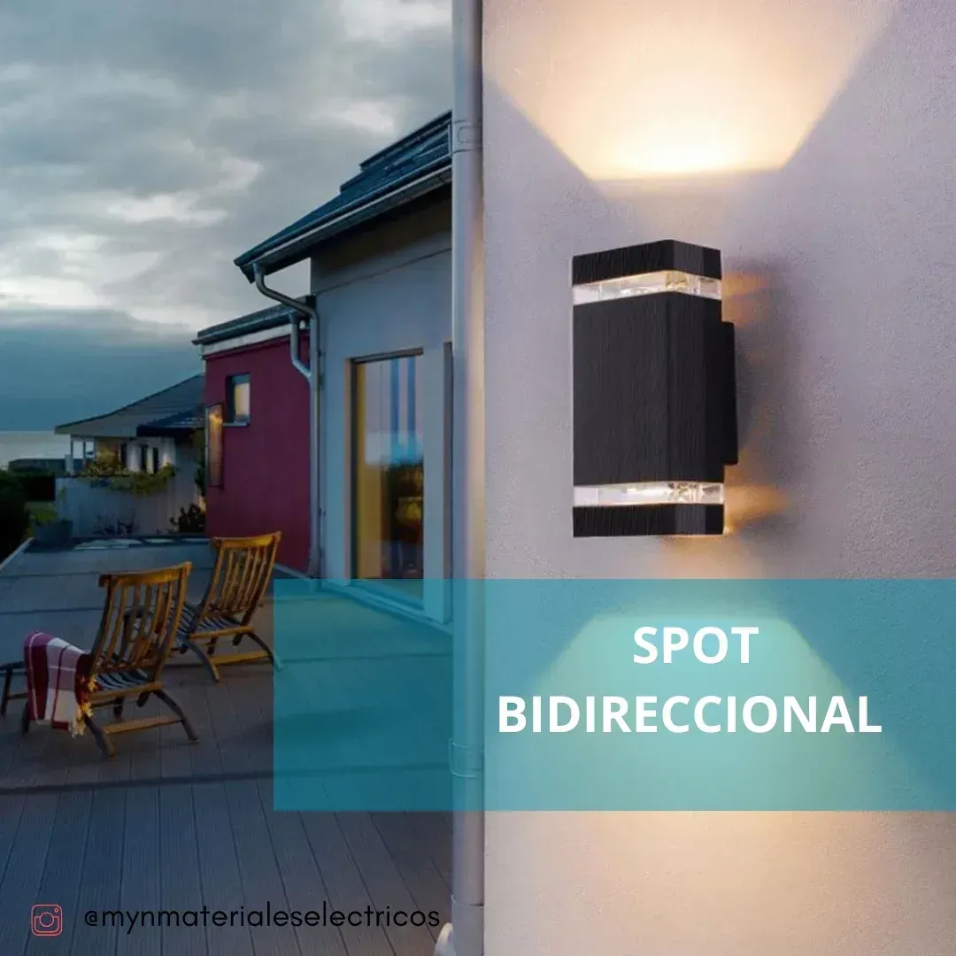 Foco bidireccional de pared negro sobre pared blanca; entorno de patio al aire libre con sillas y una casa.