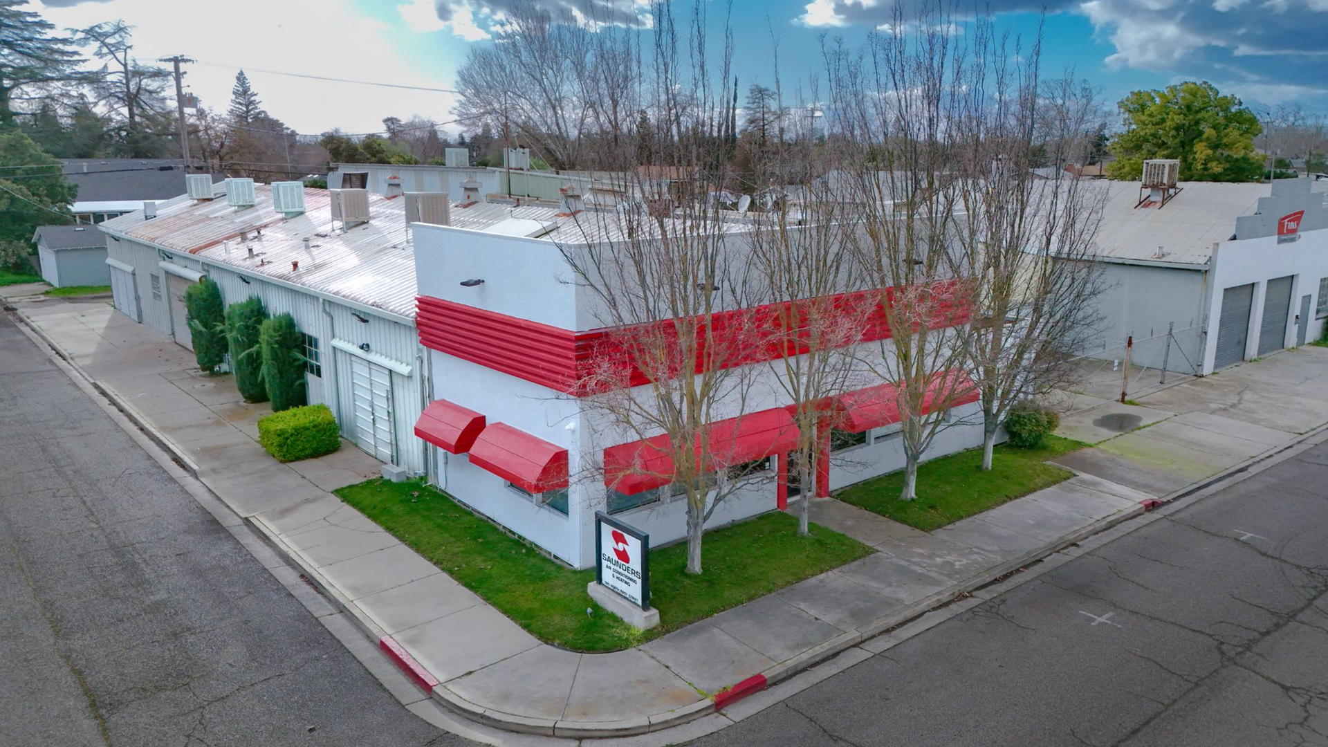 Building with red and white facade, red awnings, green grass, and trees on a corner lot under a cloudy sky.