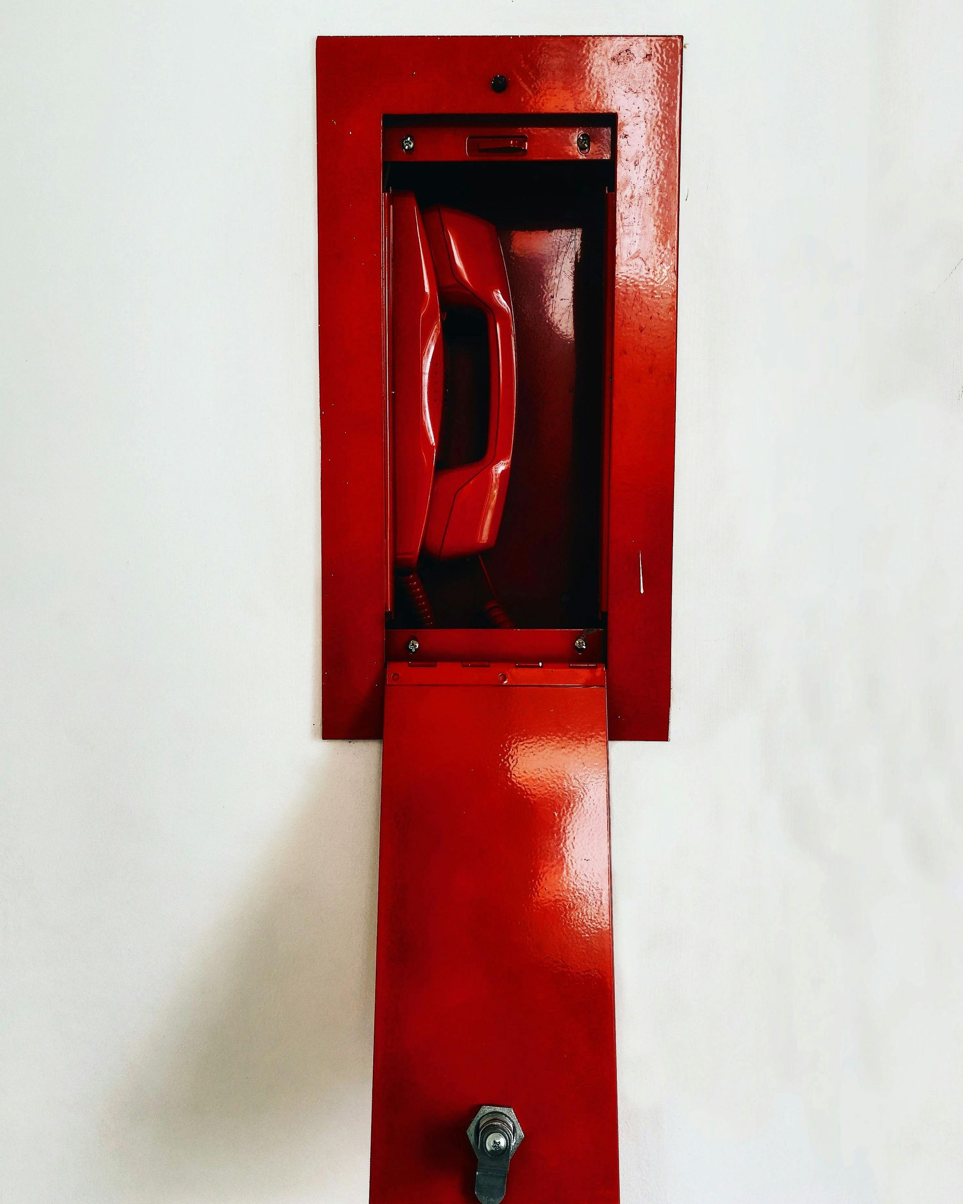 Red phone in a red metal box mounted on a white wall. The box has a key lock and a hinged door.