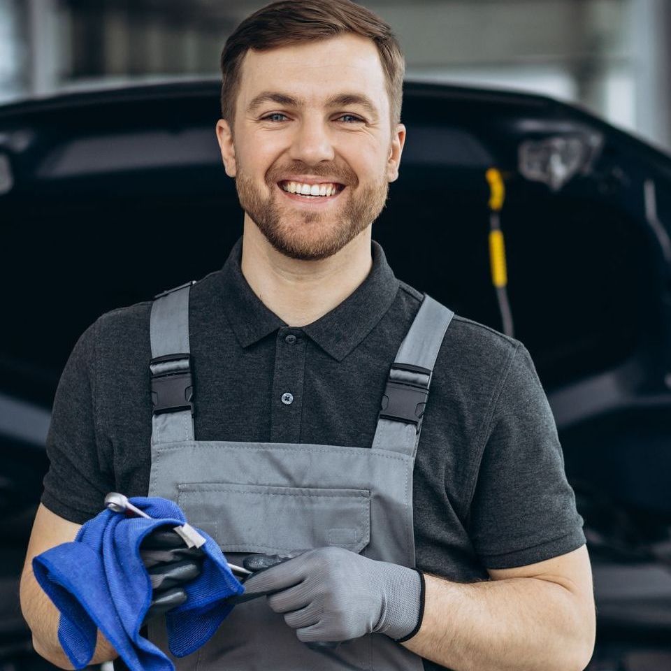 A man in overalls is holding a wrench and a towel in front of a car.