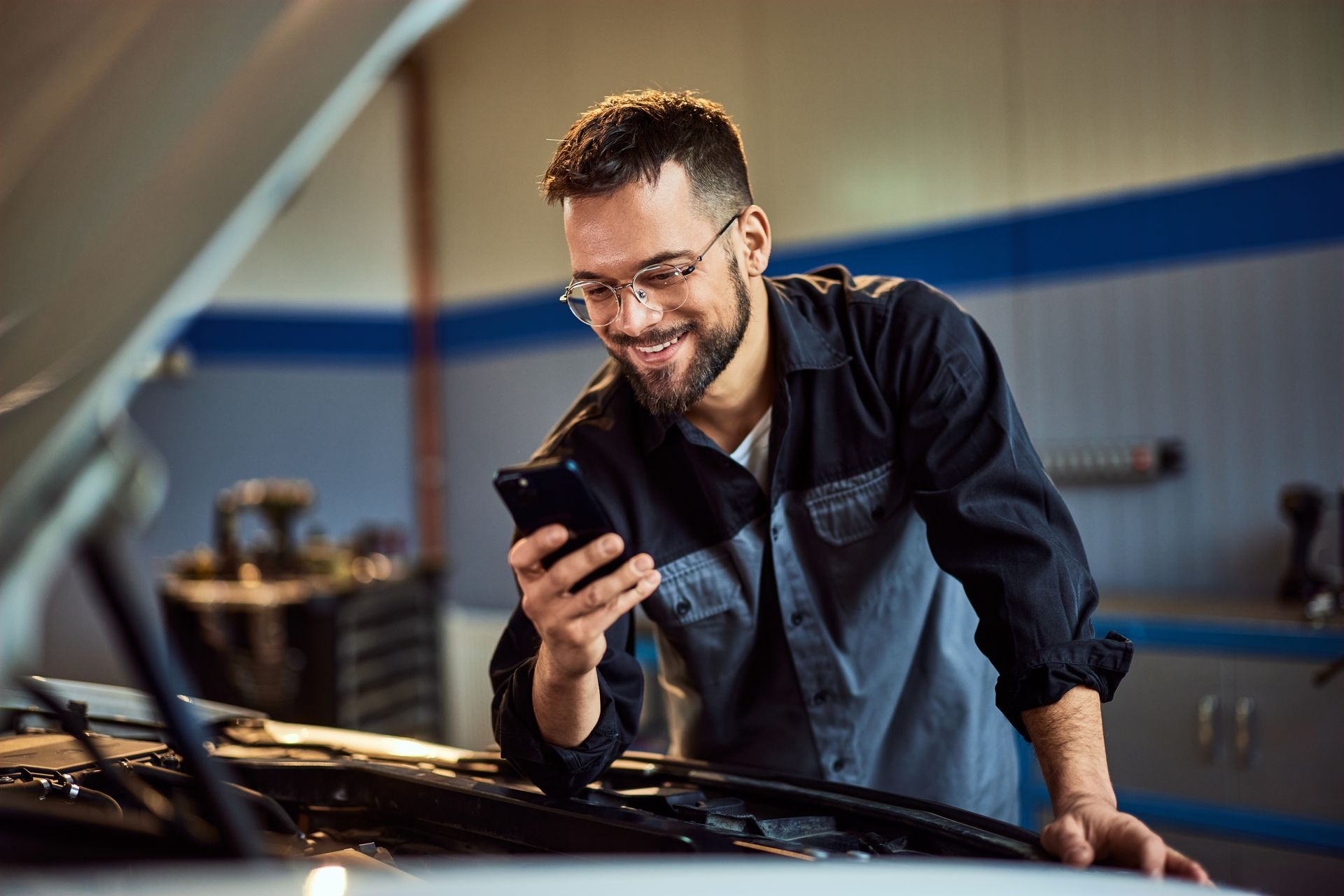 A mechanic is looking at his cell phone while standing next to a car.