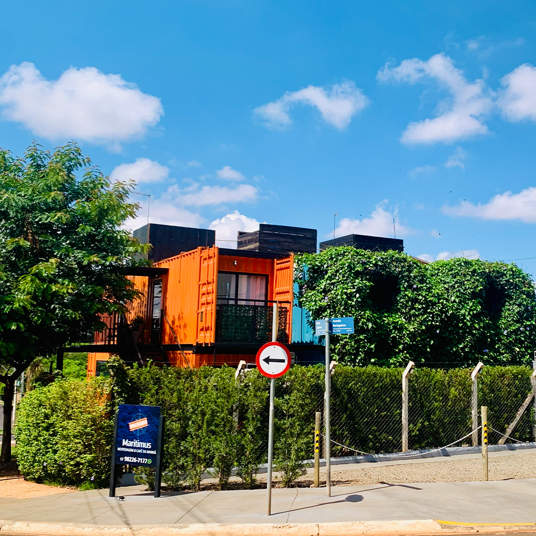 Prédio colorido feito de contêineres marítimos, com árvores, céu azul e uma placa de rua de sentido único.
