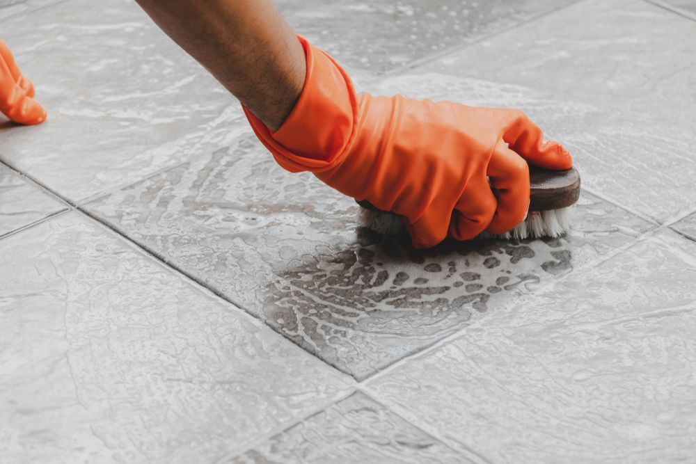 Gloved Hand Scrubbing Tiled Floor With A Brush; Soapy Water Visible — Rocky Commercial Cleaners In Berserker, QLD