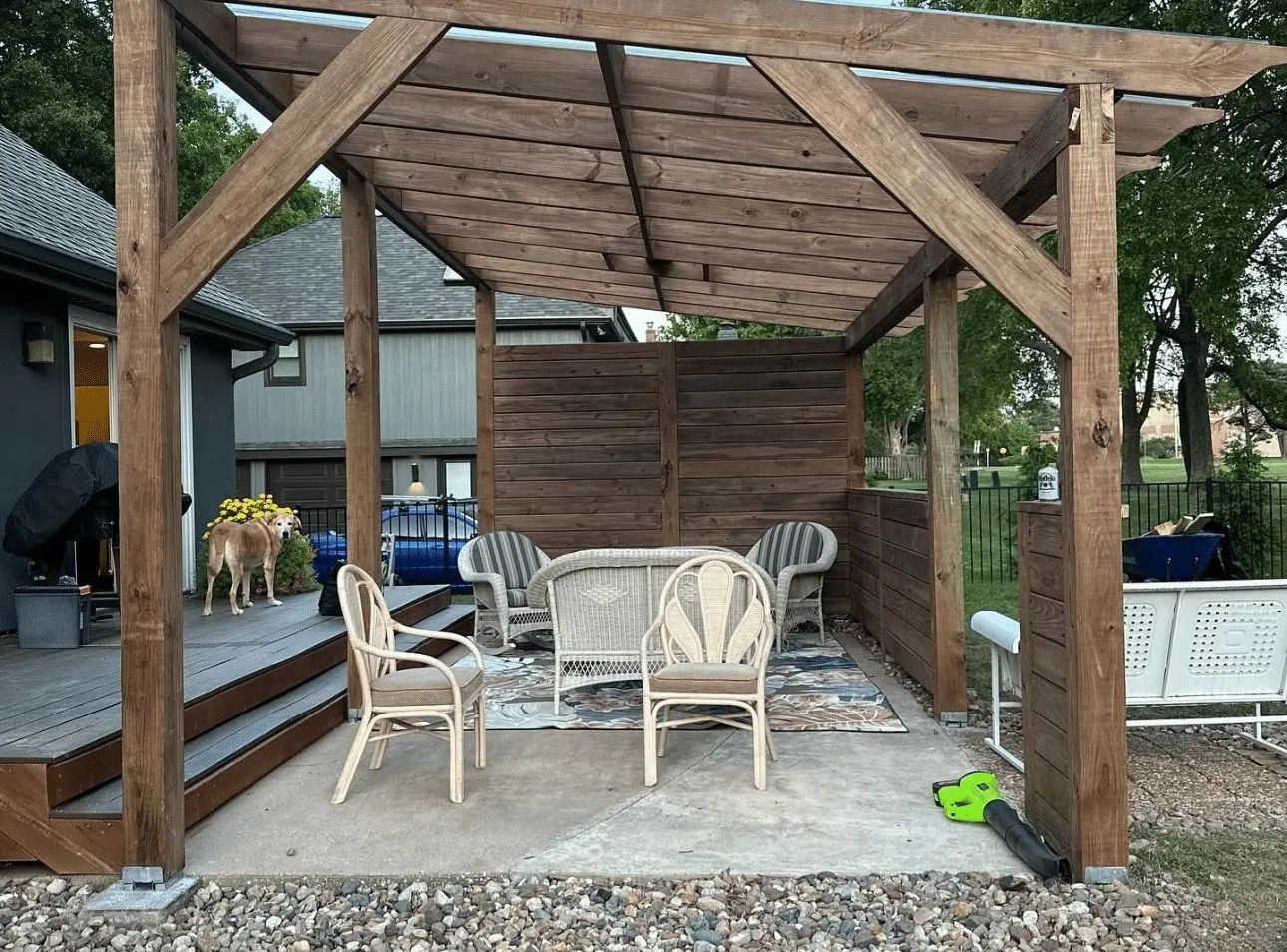 A wooden pergola covers a concrete patio with four wicker chairs and a rug, adjacent to a house with a raised wooden deck.