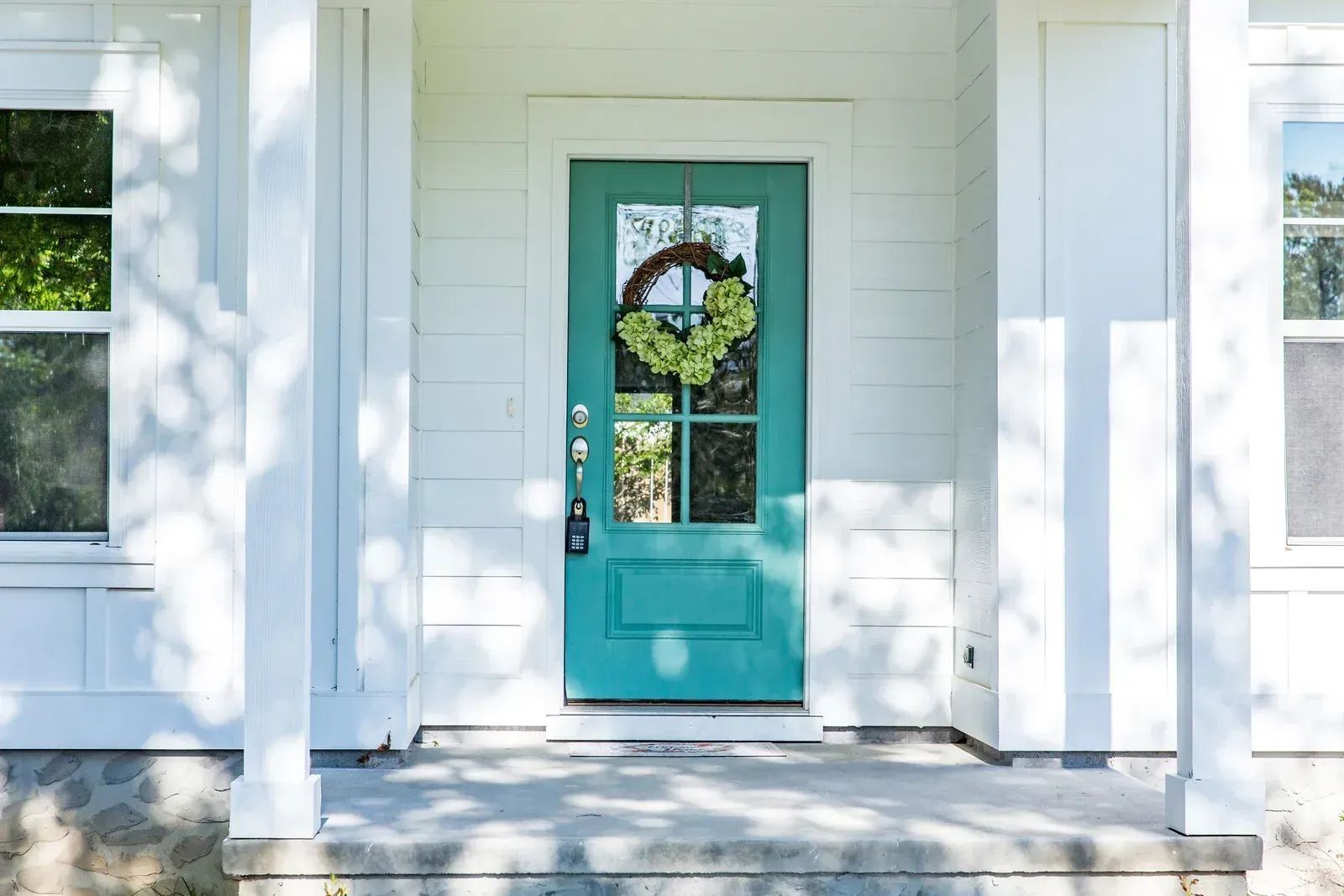 Teal front door with glass panes, green wreath, white siding, and porch under sunlight.