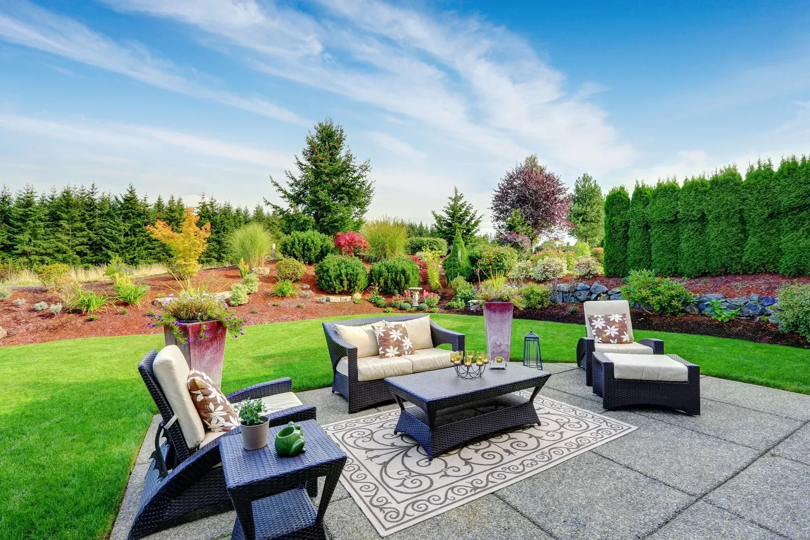 Outdoor patio with seating area on stone, green lawn, and lush garden under a blue sky.
