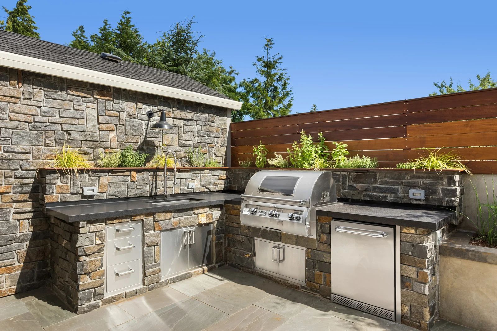Outdoor kitchen with stone walls, stainless steel grill, cabinets, and a wood fence against a blue sky.