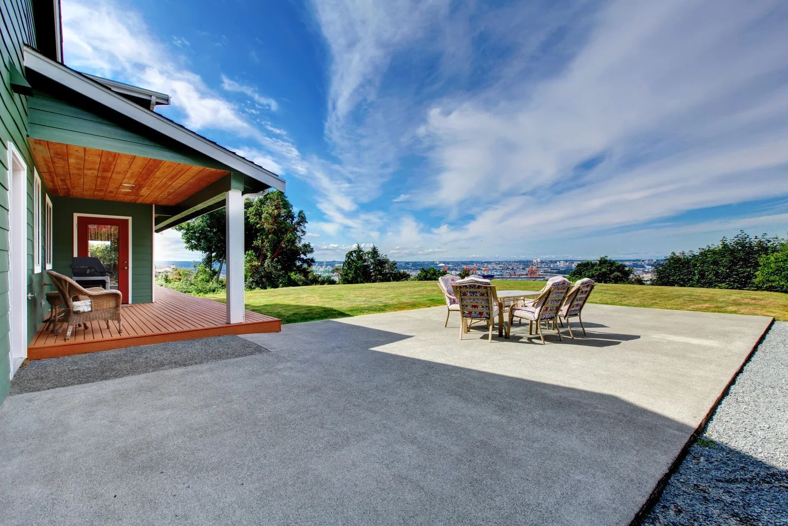 A green house with a covered brick porch and a wide concrete patio overlooking a grassy hillside under a blue sky.