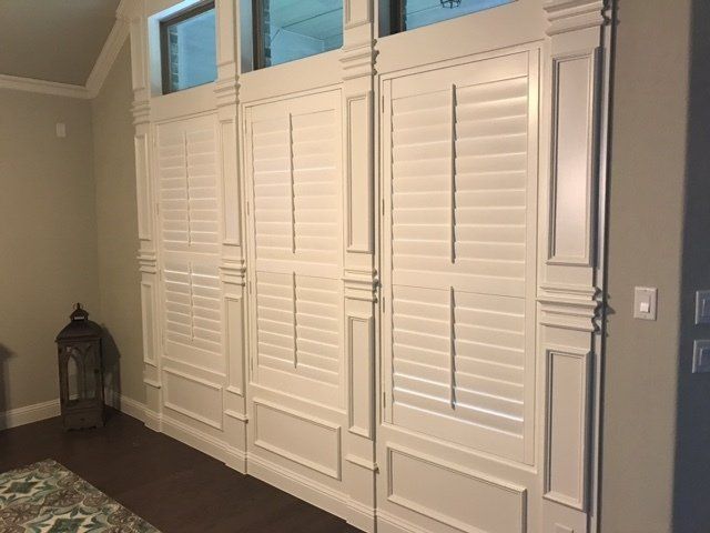 White shutters on a wall with decorative trim. Light fixture and dark wood flooring are also visible.