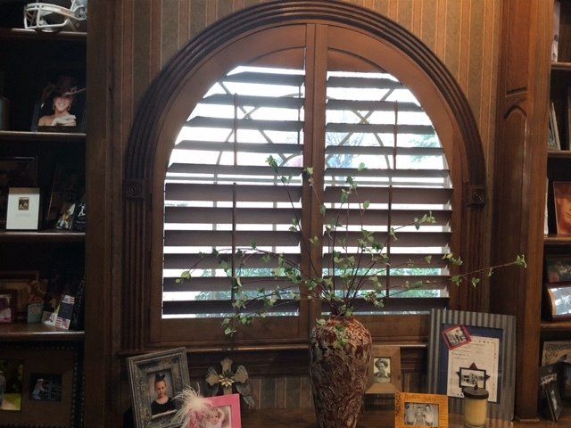 Wooden arched window with closed shutters, surrounded by bookshelves and framed photos.