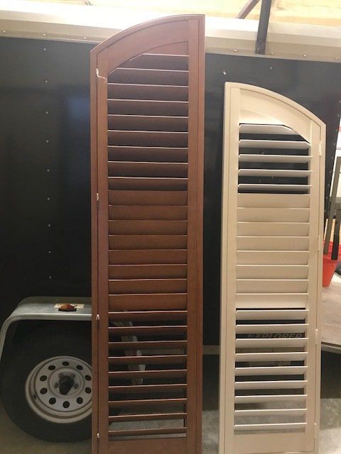 Brown and cream-colored louvered shutters propped against a black trailer. The left one is wood, the right one is painted.