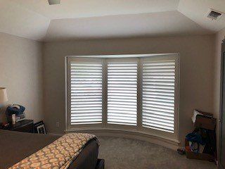 Bedroom with bay window and white shutters. Neutral walls, ceiling with a recessed area, and a bed visible.