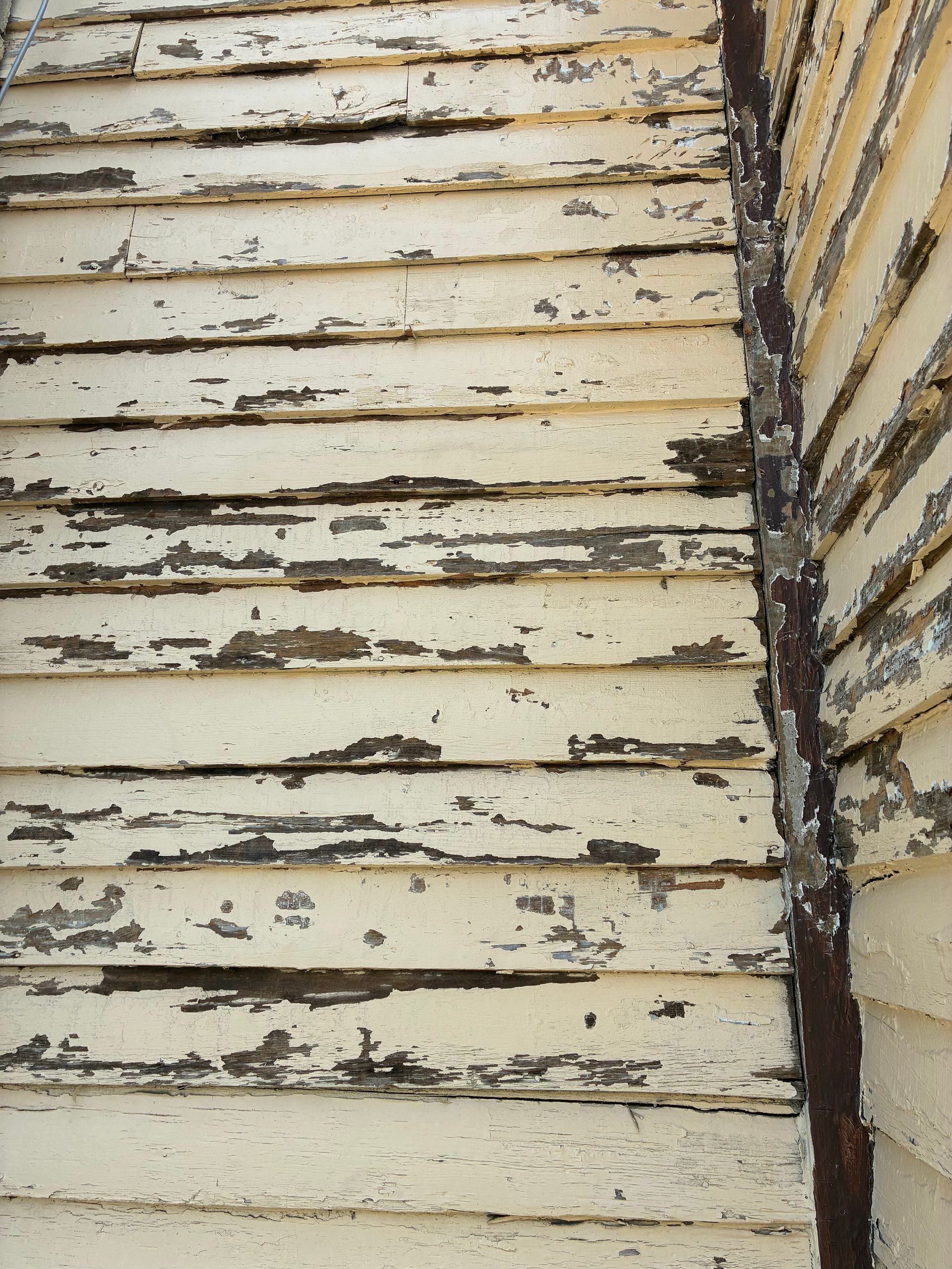 Weathered, peeling cream-colored siding on the corner of a building. A rusty metal corner piece is visible.