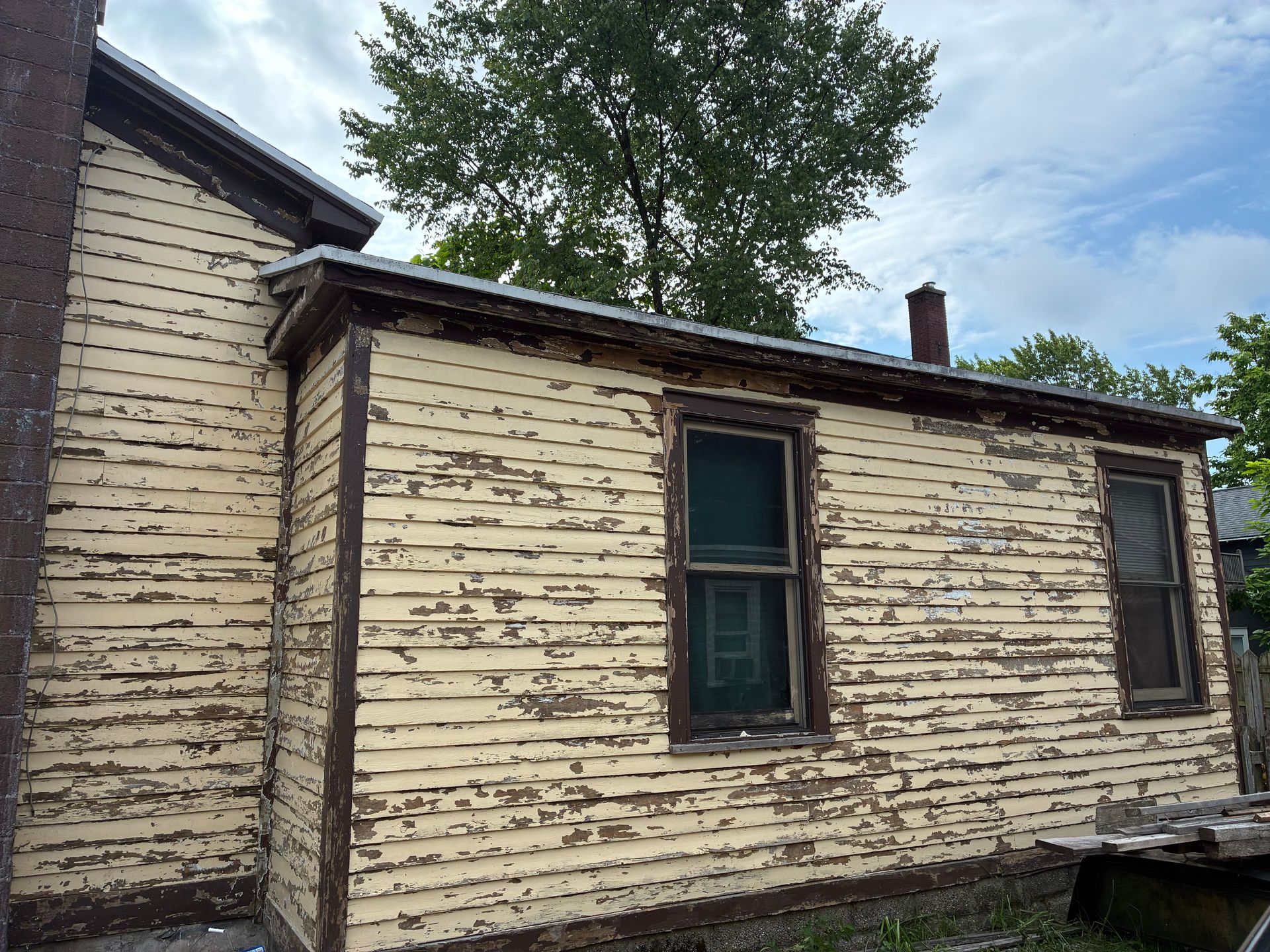 Side view of a weathered yellow house with peeling paint, two windows, and a chimney.