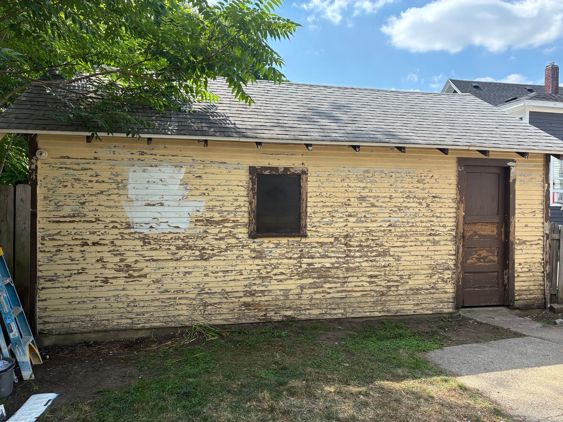 Weathered yellow garage with peeling paint, brown door, small window, and gray roof under a blue sky.