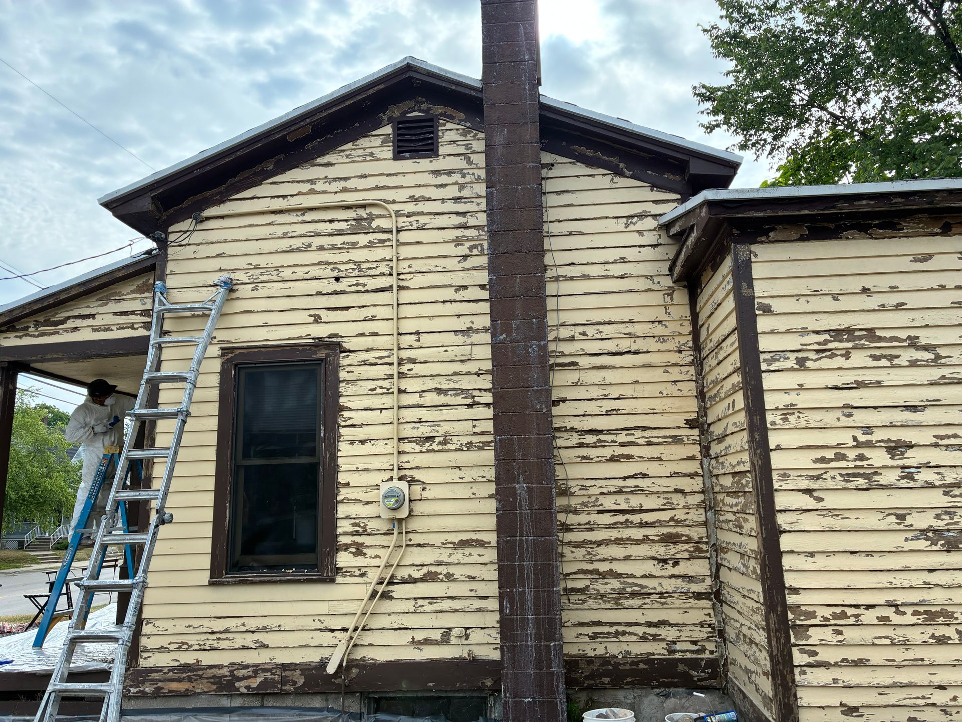 A person on a ladder is scraping peeling yellow paint from a house exterior.