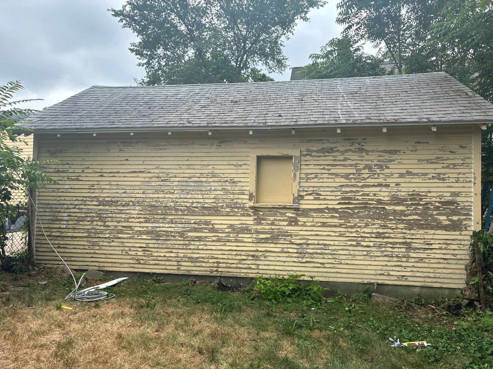 Weathered, yellow shed with peeling paint, overgrown grass, and a boarded-up window.