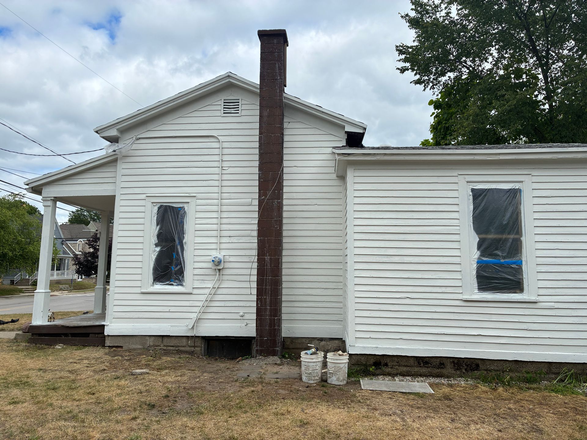 White house with brown chimney and covered windows; buckets on the ground, overcast sky.