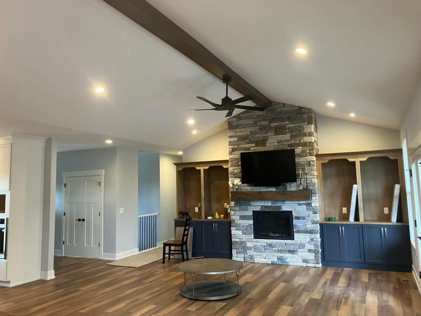 Living room with stone fireplace, TV, built-in cabinets, wood floors, and a ceiling fan.