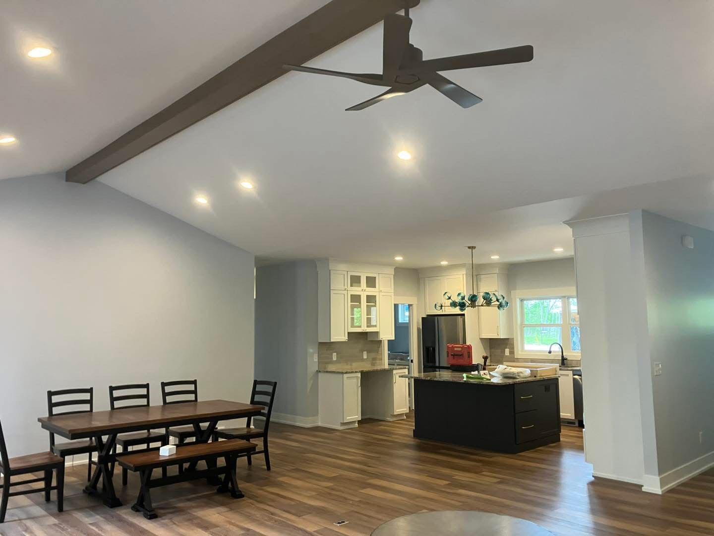 Open-plan dining room and kitchen. Dark wood table, black chairs. Kitchen with white cabinets, black island, and brown ceiling beam.