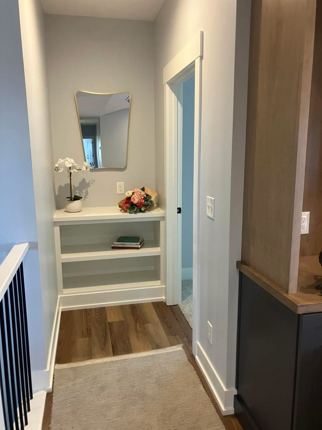 Hallway with a shelf, mirror, flowers, and door. Light gray walls, wood floor, and a neutral rug.
