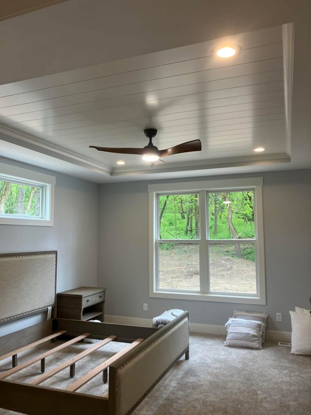 Bedroom with tray ceiling, ceiling fan, light blue walls, window with view of trees, and bed frame.