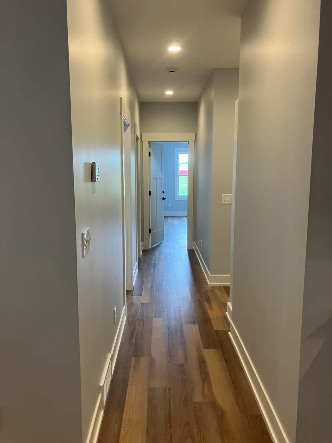 Narrow hallway with light gray walls, wood-look floor, and recessed lighting. A door is at the end.