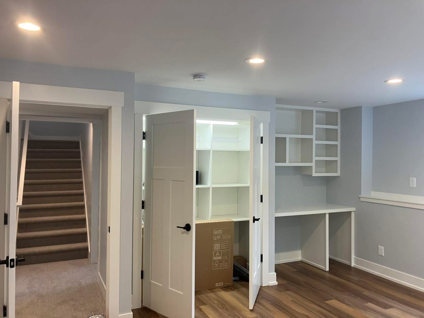 A basement room with a built-in desk and shelving. Stairs are visible to the left, a white closet with shelves is open.