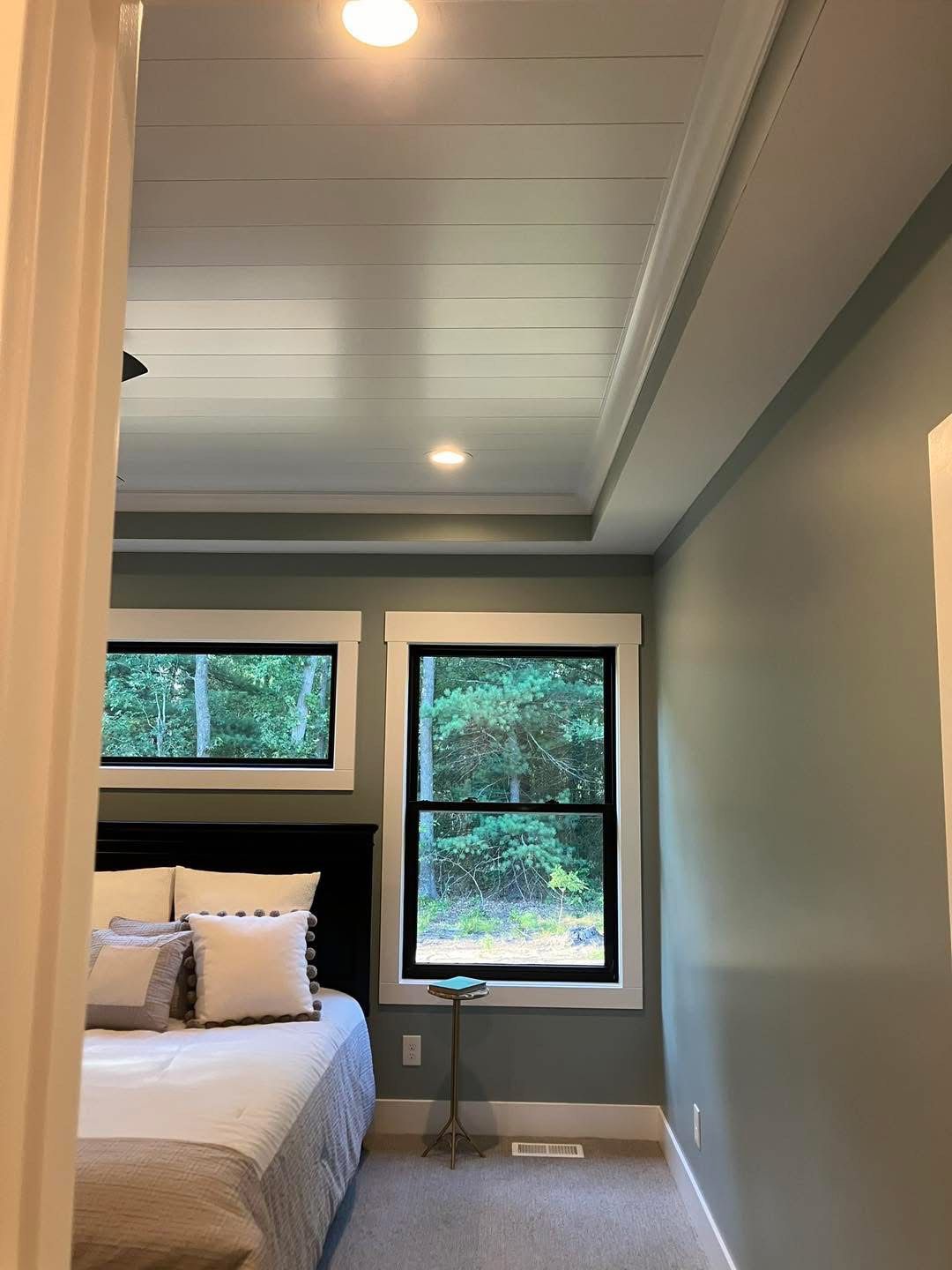 Bedroom with blue-green walls, white trim, plank ceiling, two windows with black frames, and a bed.