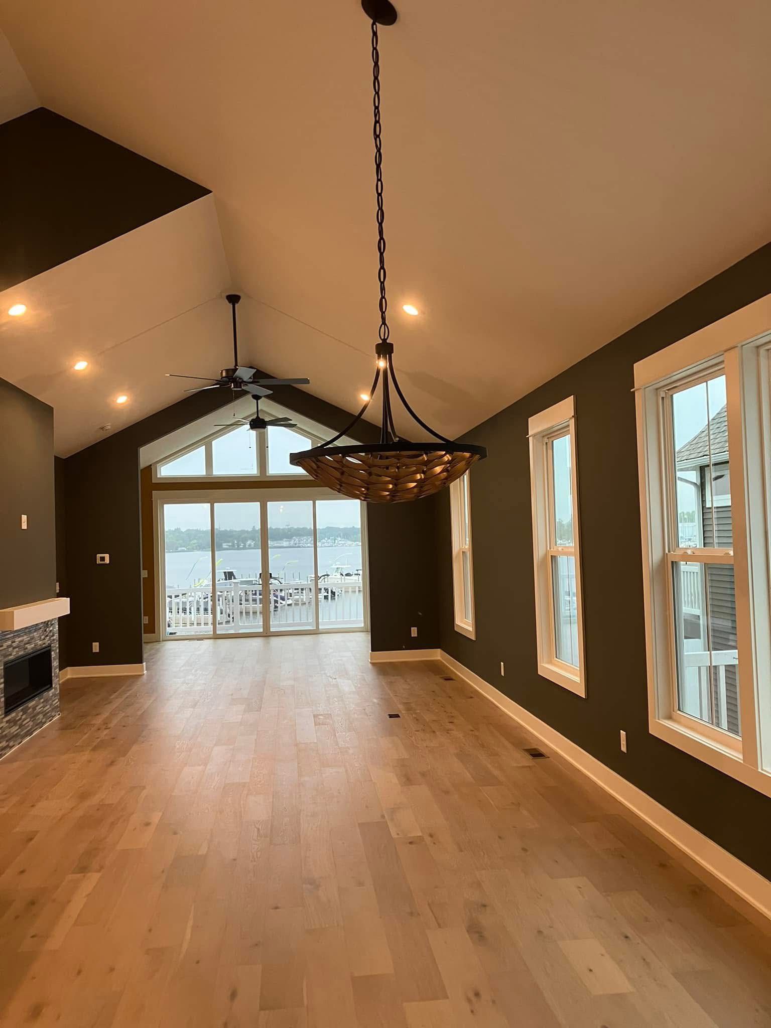 A spacious living room with hardwood floors, dark green walls, and large windows overlooking a waterfront.