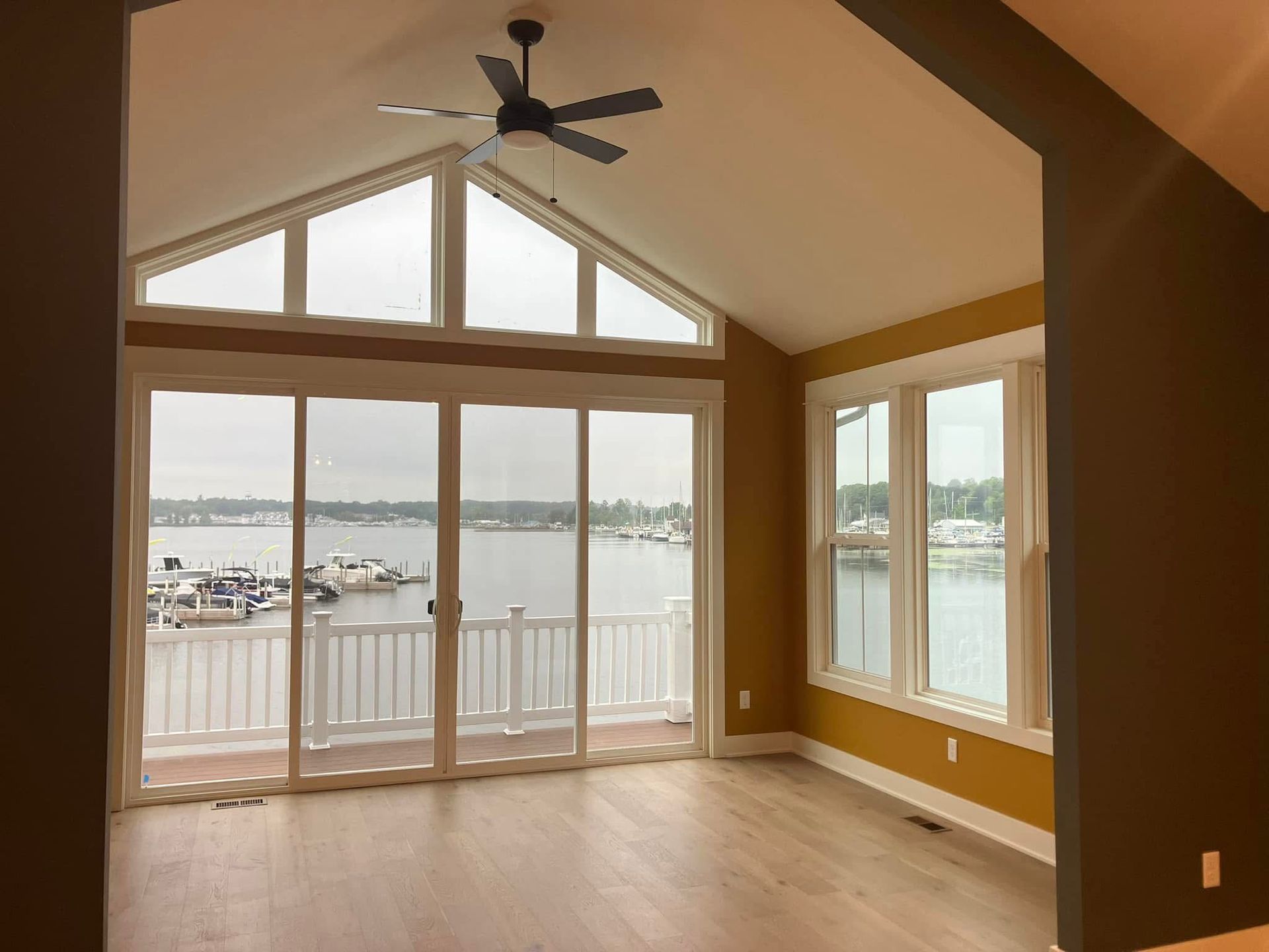 Bright room with large windows overlooking a marina. Light wood floors, mustard walls, and a dark ceiling fan.