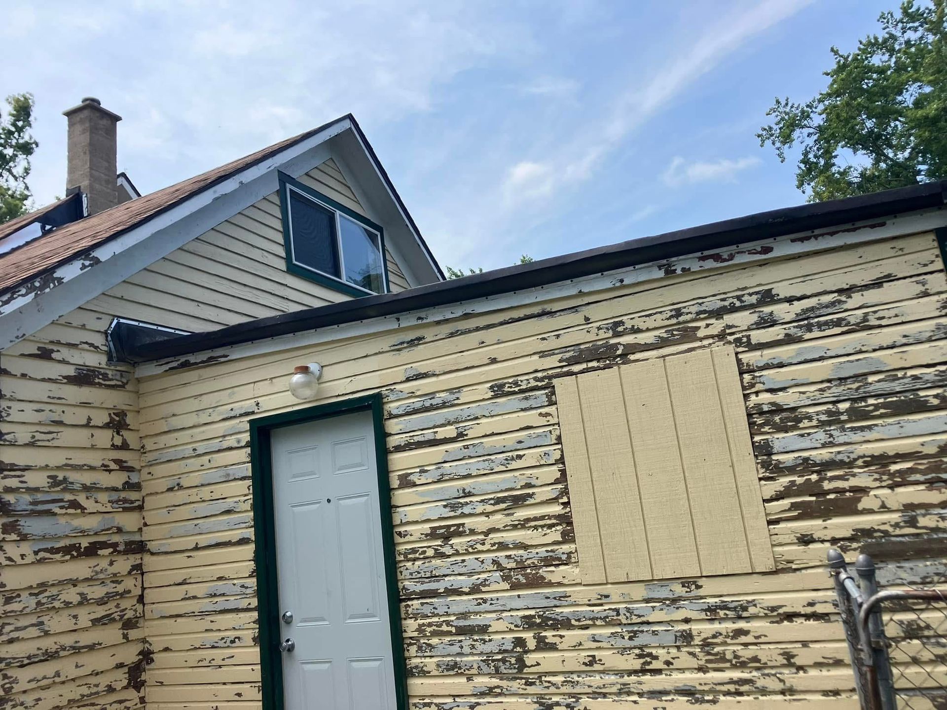 Yellow house with peeling paint, black trim, white door, under a blue sky.