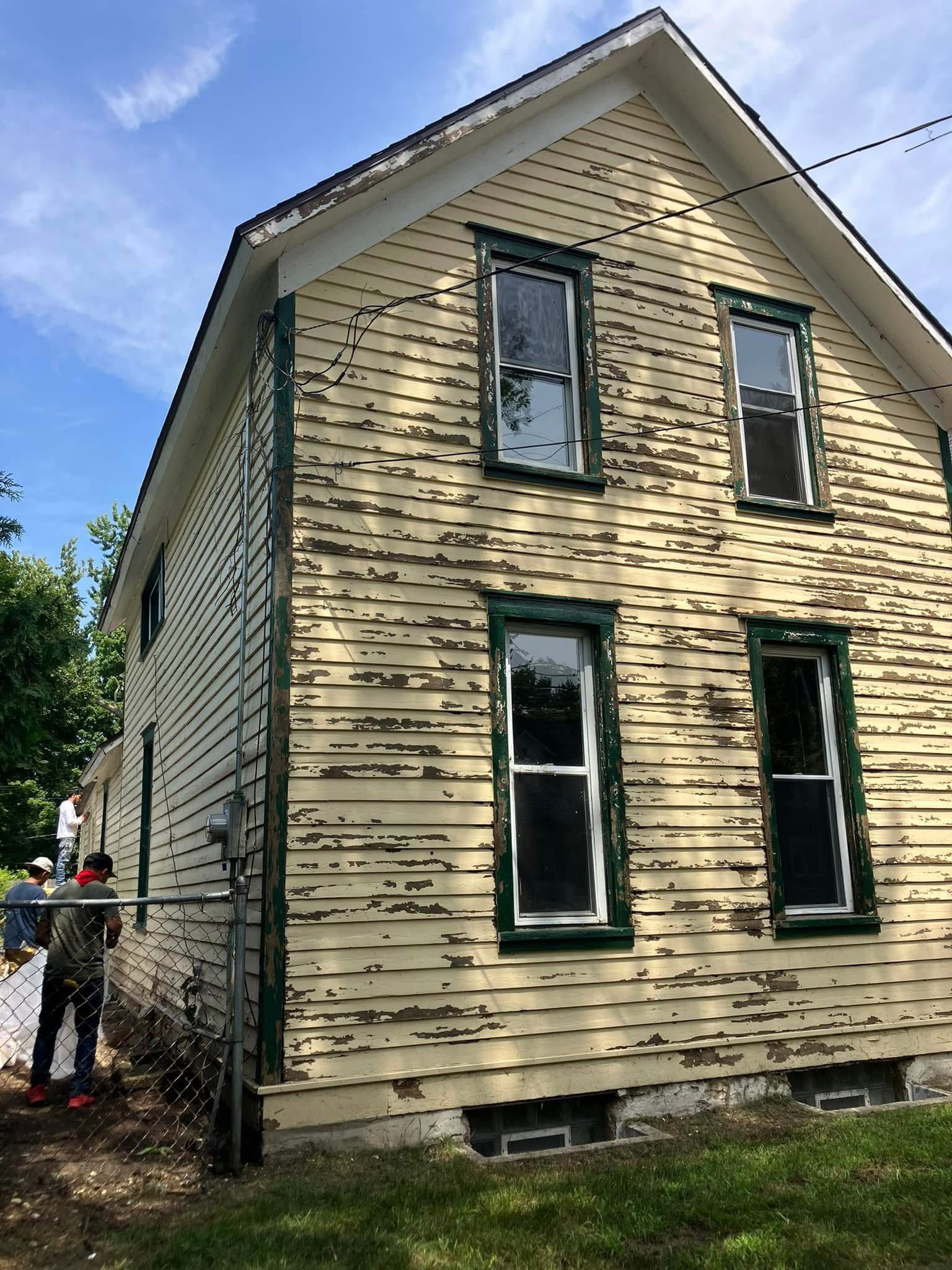 Two-story yellow house with peeling paint, green trim, and visible windows. A person stands nearby, fence in front.
