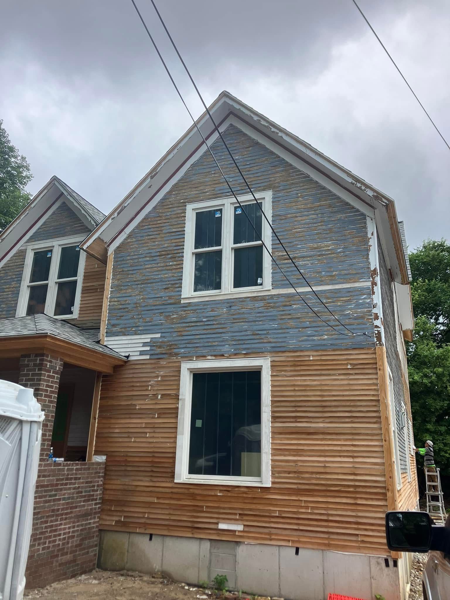 Two-story house under construction, exposed wood siding and blue-gray shingle siding, gray sky.
