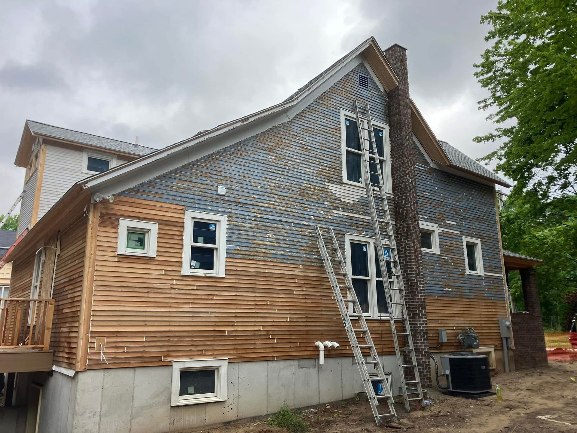 Side view of a house undergoing construction; siding being removed, ladder against the wall, cloudy sky.