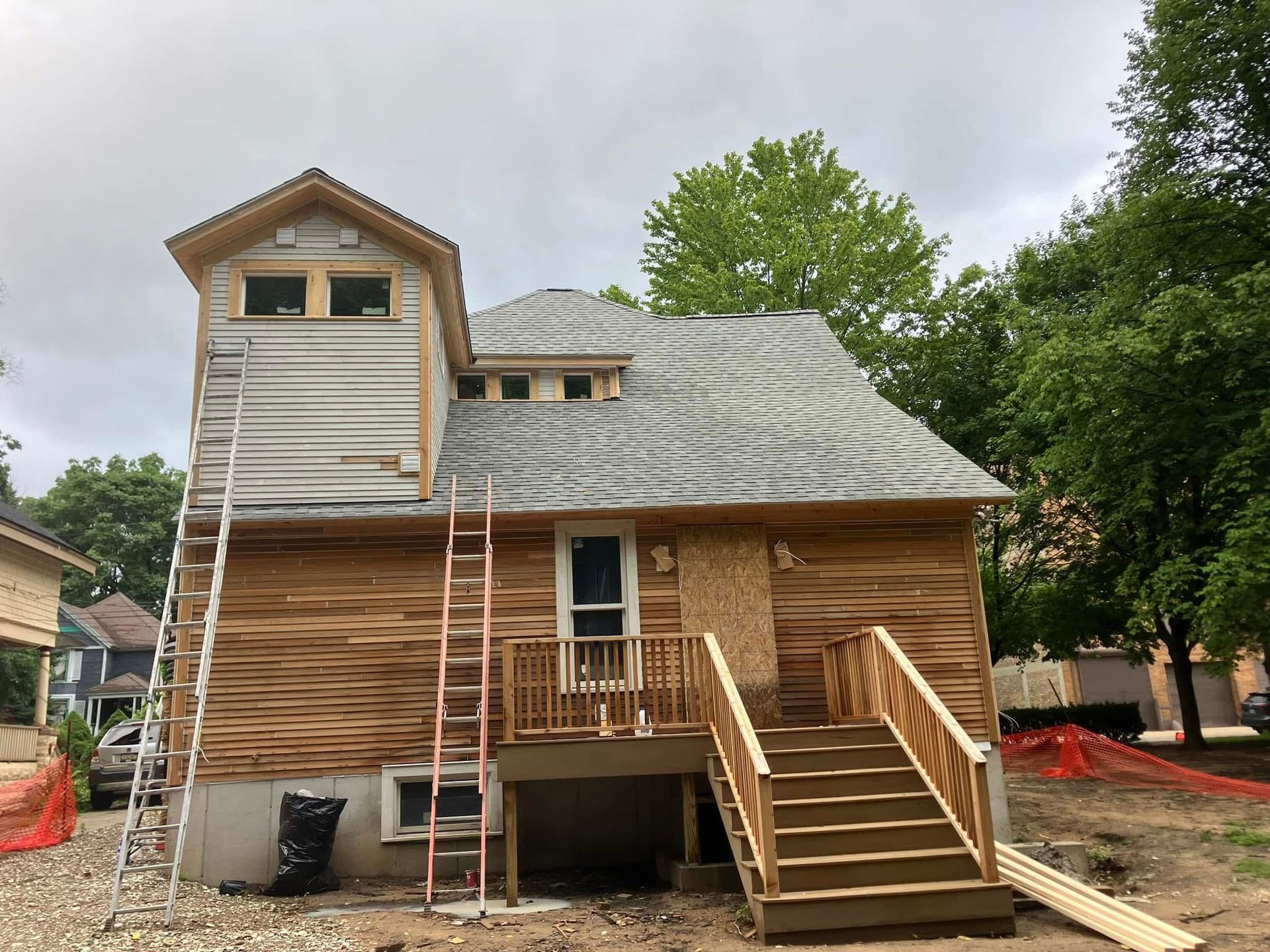 House under construction with wooden siding, stairs, and ladder. Gray roof with dormer windows.