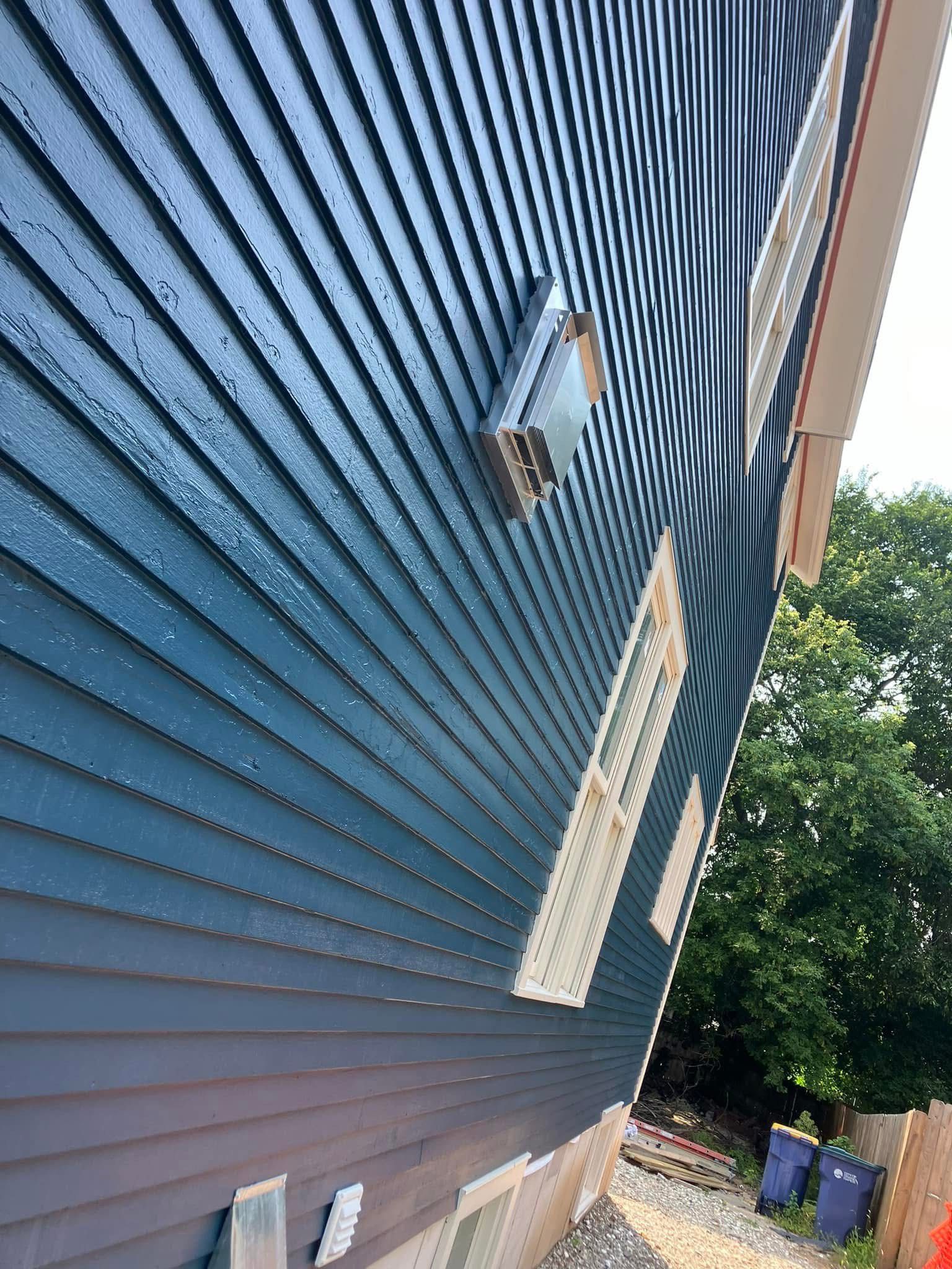 Blue clapboard siding on a building exterior, with windows and a vent, with a treeline visible in the background.