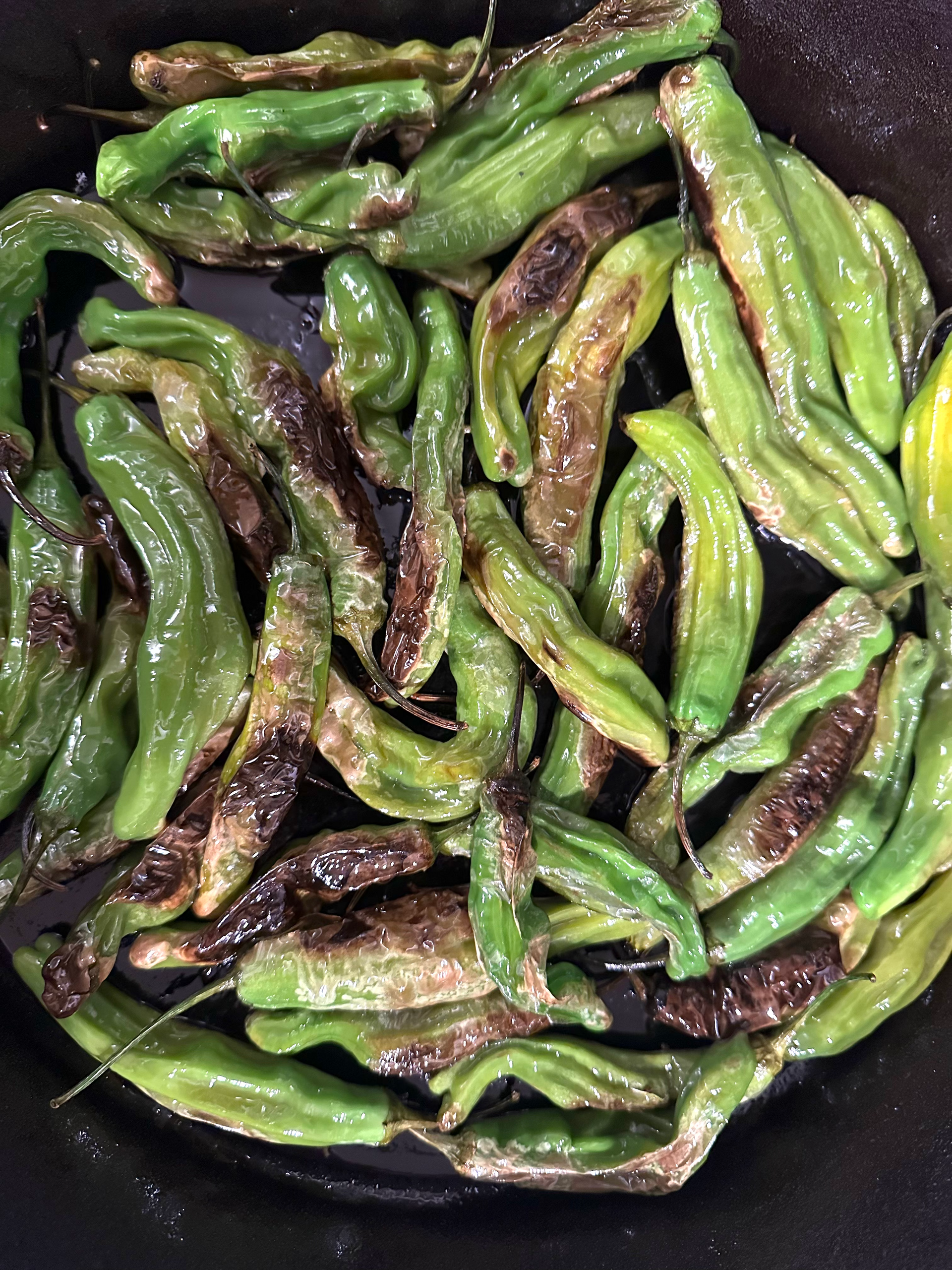 Sautéed green shishito peppers in a dark pan, some blistered and charred.