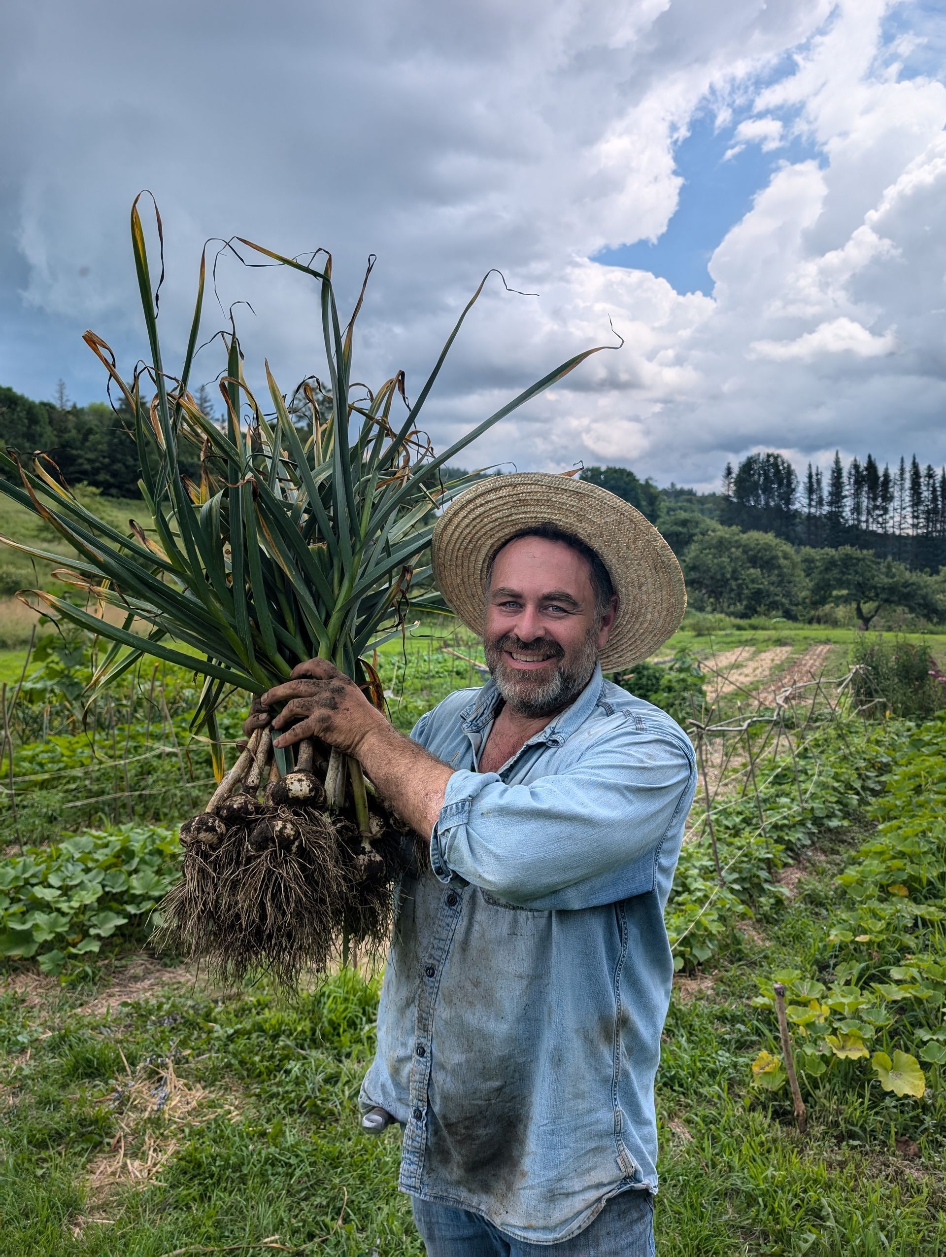 Farmer in straw hat smiles, holding harvested onions in a field under cloudy sky.
