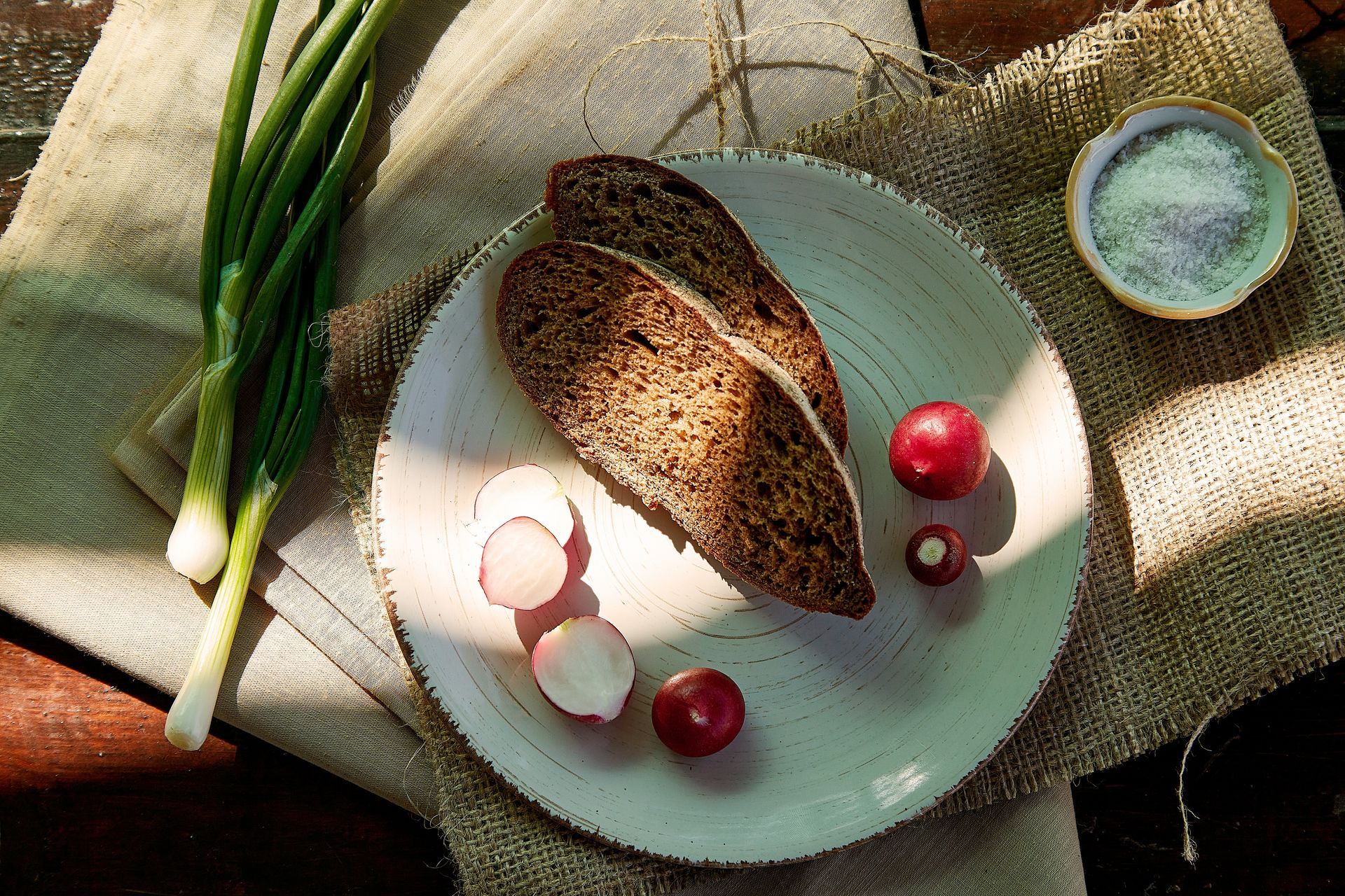 Sliced bread, radishes, and green onions on a plate, with salt and burlap, lit by sunlight.