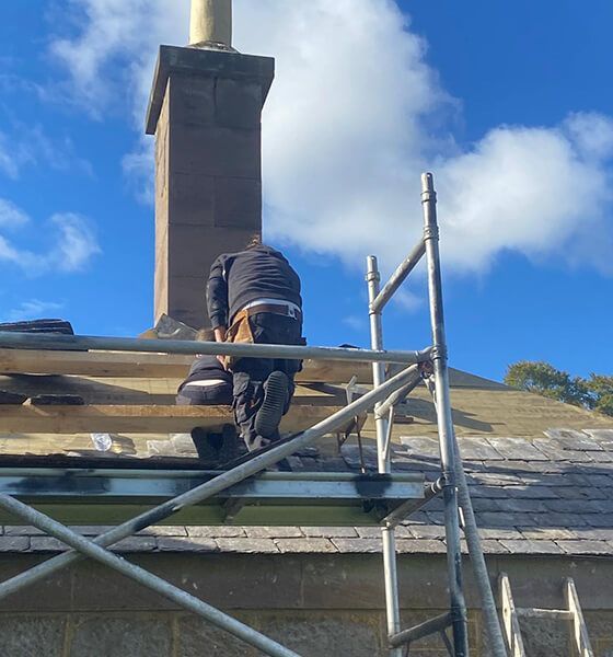 Workers on scaffolding near a chimney repair a roof on a sunny day.