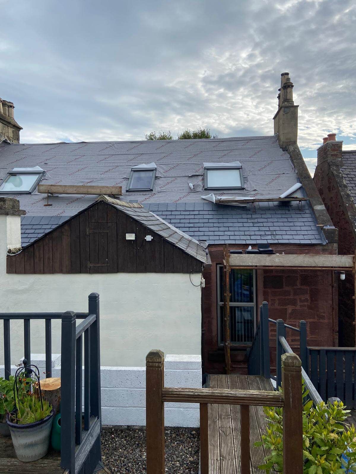 A slate roof with three skylights, supported by wooden beams, over a white and brown building; overcast sky.
