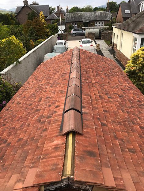 Overhead view of a red tiled roof with a central ridge. Houses and trees in the background.