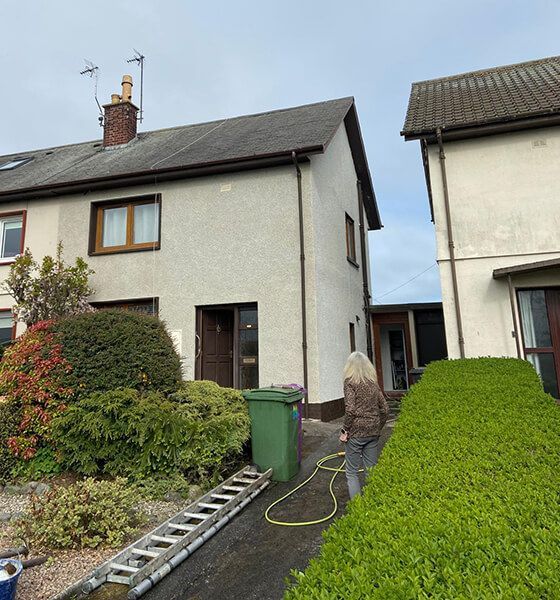 Two-story house with green bin, hedge, and a person walking with a hose.