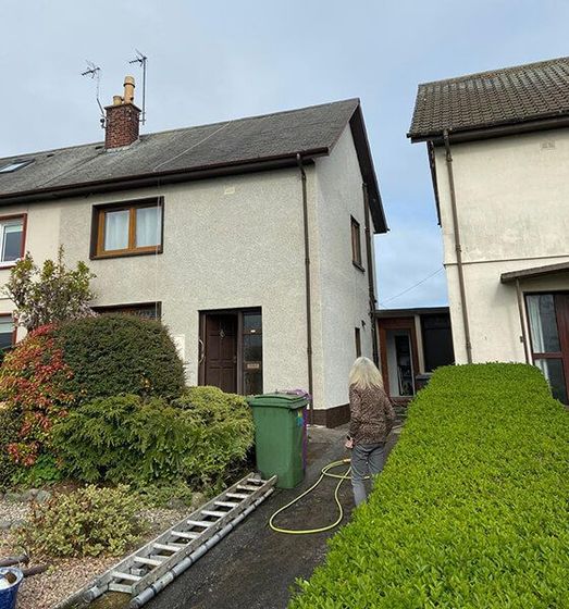 Two-story house with green bin, hedge, and a person walking with a hose.