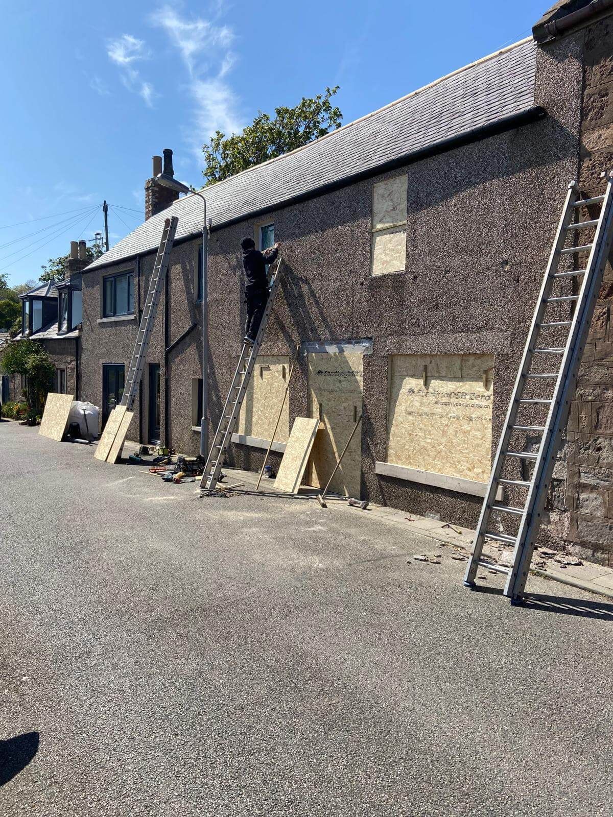 Man on ladder working on boarded-up brick building exterior; ladders, plywood, and debris visible.